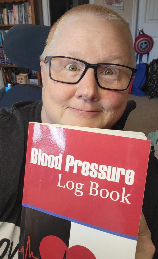 Spoonie-friendly products can include things like the Blood Pressure Log Book that Janni is holding up in this photo. She is looking at the camera and smiling. 