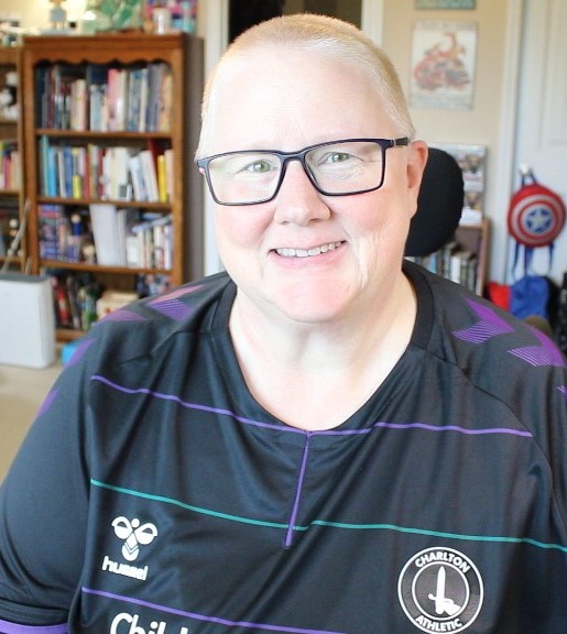 Chronic Illness Spoonie Janni .She has a very short, buzzed off hairstyle. She is wearing eyeglasses and smiling at the camera. She's wearing a black jersey representing her beloved Charlton Athletics football team. 